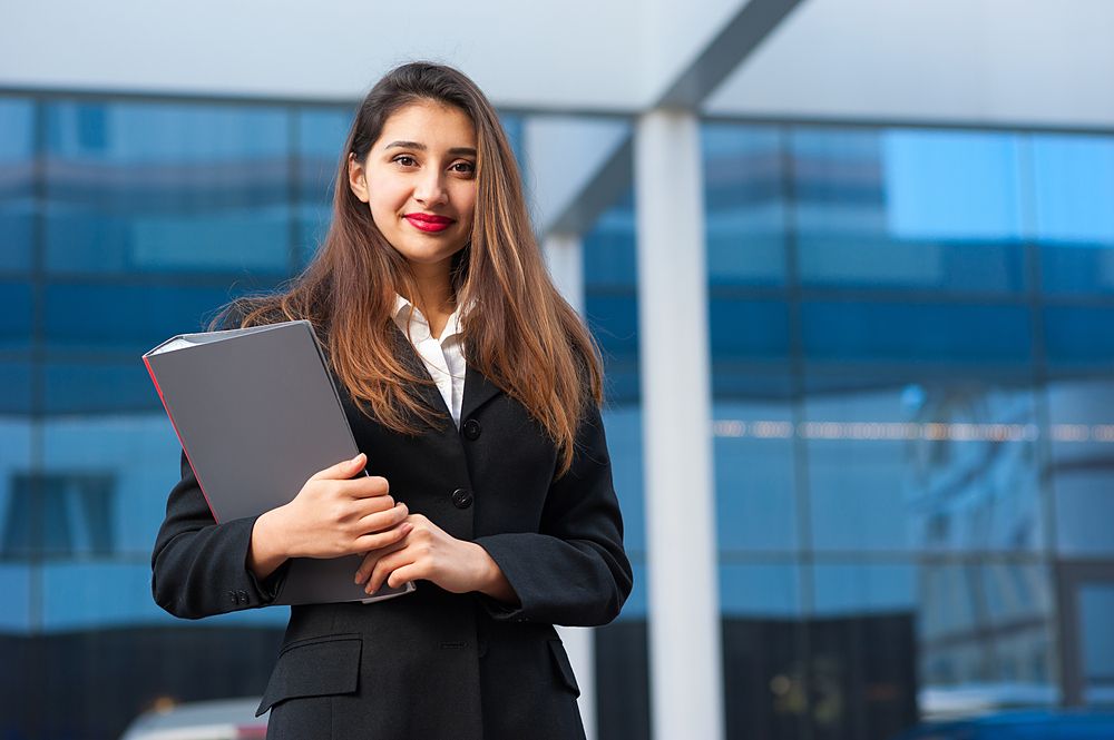 A businesswoman in a black suit holding a folder outside an office.
